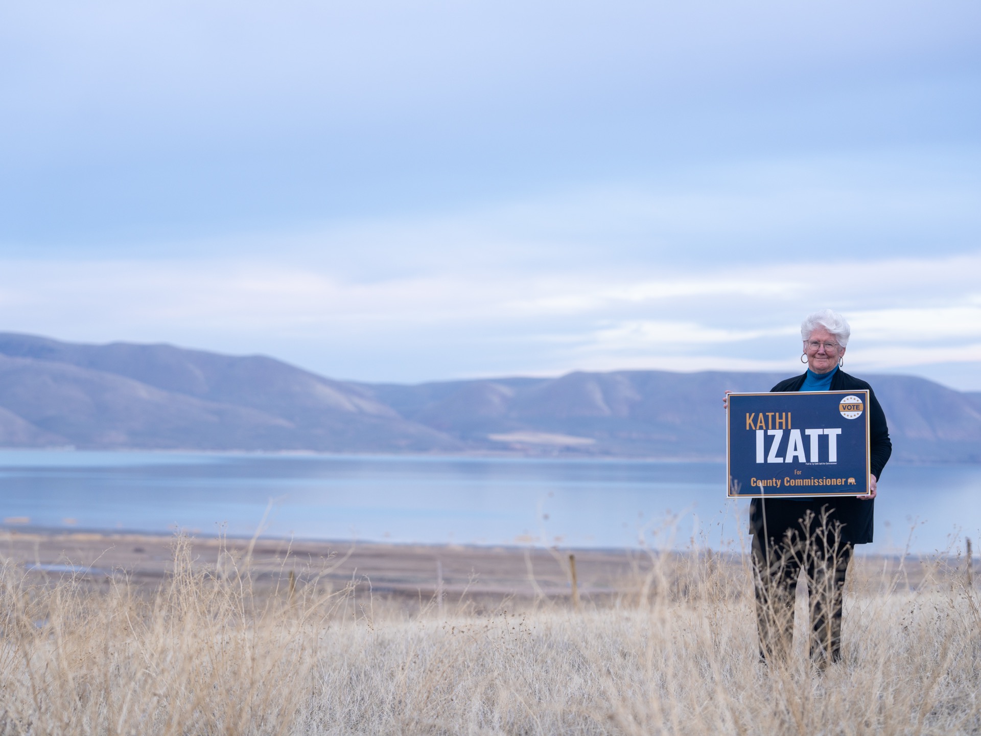 Kathi Izatt standing in a field overlooking Bear Lake, holding her campaign sign